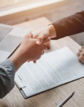 Two people are shaking hands over a document on a clipboard, marking the beginning of a collaborative journey. A pen is placed nearby on the wooden table, poised for contributions from their peer group.