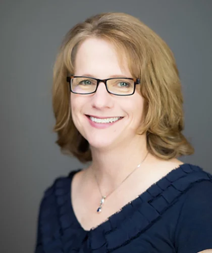 A person with shoulder-length light brown hair, wearing glasses and a navy blue top, smiles warmly amid their peer group against a gray background.