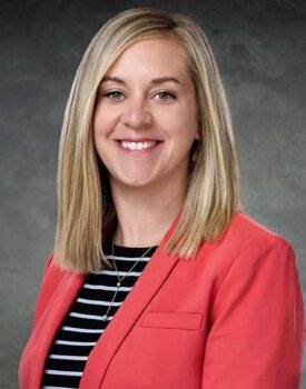 Amanda Huffman, a woman with straight blonde hair, smiles in front of a gray background while wearing a coral blazer over a black and white striped shirt.