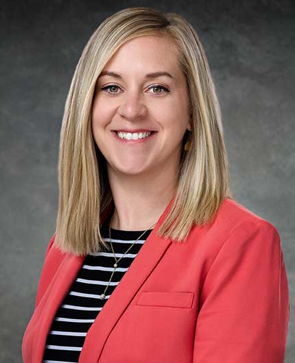 Amanda Huffman, a woman with straight blonde hair, smiles in front of a gray background while wearing a coral blazer over a black and white striped shirt.