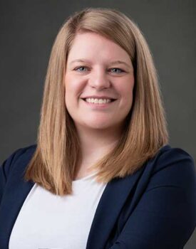 Amanda Huffman, with straight, shoulder-length blonde hair, smiles at the camera. She is wearing a navy blazer over a white top against a plain dark background.