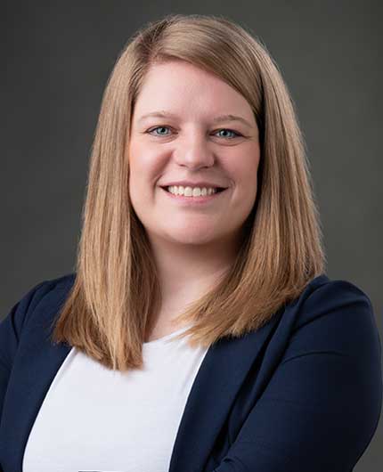 Amanda Huffman, with straight, shoulder-length blonde hair, smiles at the camera. She is wearing a navy blazer over a white top against a plain dark background.