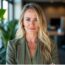 Jessica Stimmel, a woman with long blonde hair, stands confidently in an office setting with plants and large windows, dressed in a stylish green blazer.