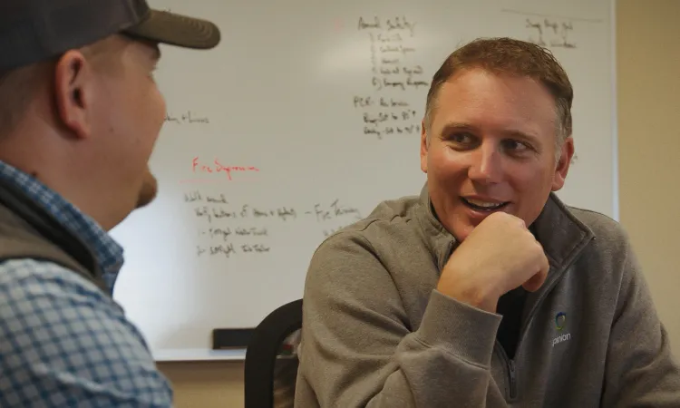 In an office setting, two men engaged in conversation, surrounded by notes on a whiteboard. Nearby, equipment dealers discussed the latest market trends.