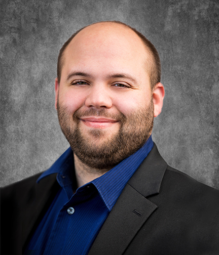 A man with a beard and short hair, wearing a black blazer over a blue shirt, poses in front of a textured gray background, exuding the confidence of a professional in the manufacturing industry.