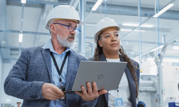 Two professionals wearing safety helmets work in a manufacturing setting. One holds a laptop, and they both appear focused on their surroundings.