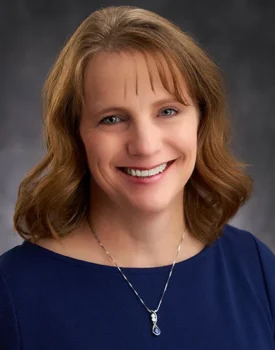 Devon with shoulder-length brown hair smiles at the camera, embodying the warmth and approachability often associated with family business advising. They wear a navy top and a necklace with a blue pendant, all set against a gray background.