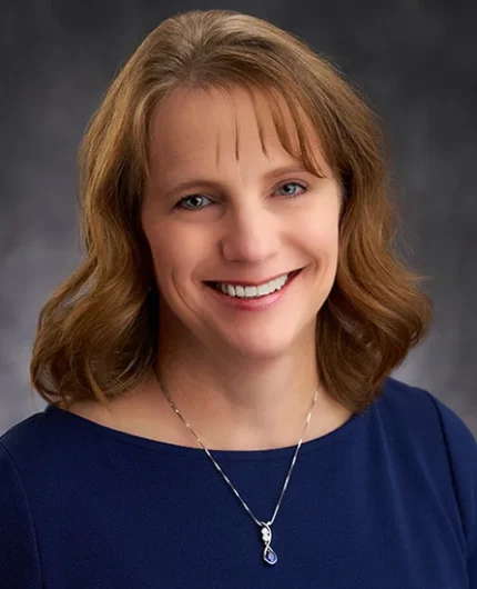 Devon with shoulder-length brown hair smiles at the camera, embodying the warmth and approachability often associated with family business advising. They wear a navy top and a necklace with a blue pendant, all set against a gray background.