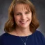 Devon with shoulder-length brown hair smiles at the camera, embodying the warmth and approachability often associated with family business advising. They wear a navy top and a necklace with a blue pendant, all set against a gray background.