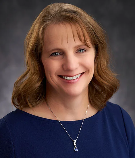 Devon with shoulder-length brown hair smiles at the camera, embodying the warmth and approachability often associated with family business advising. They wear a navy top and a necklace with a blue pendant, all set against a gray background.