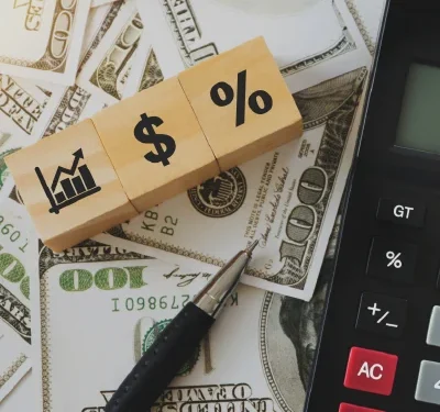 A calculator and pen rest atop various dollar bills, alongside three wooden blocks featuring financial symbols: a bar chart, a dollar sign, and a percentage sign—tools essential for any family business advising session.