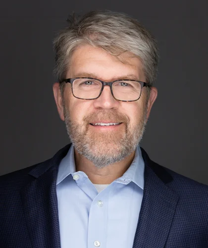 Lance Woodbury, a bearded man with glasses, smiles in a professional headshot, donning a blue shirt and dark jacket against a plain gray background.