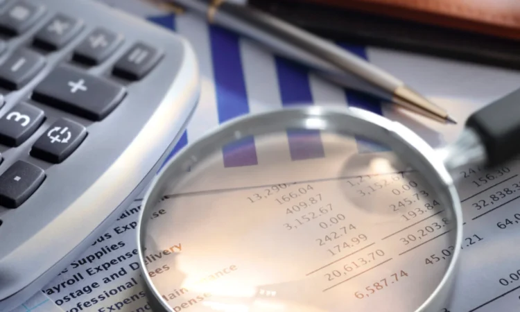 A magnifying glass hovers over financial documents, revealing numbers under scrutiny, with a calculator, pen, and bar graph in the background—a visual echo of a meticulous audit process.