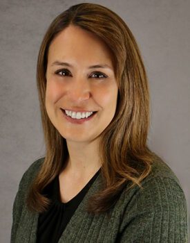 Amy Forsman, a woman with straight light brown hair and a green sweater, smiles at the camera in front of a neutral gray background.