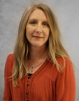 Erin Furr, a woman with long blonde hair, wears a rust-colored blouse and a necklace as she poses in front of a gray background.