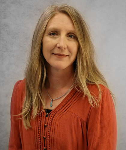 Erin Furr, a woman with long blonde hair, wears a rust-colored blouse and a necklace as she poses in front of a gray background.