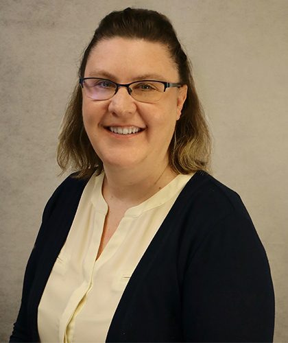 Sarah Stanger, a woman with shoulder-length brown hair, wearing glasses, a white blouse, and a dark cardigan, smiles at the camera against a plain light background.