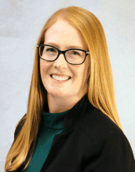 Erin Stockwell, a woman with long red hair and glasses, smiles warmly in a dark blazer and green top against a light neutral background.