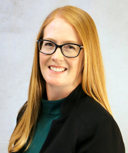 Erin Stockwell, a woman with long red hair and glasses, smiles warmly in a dark blazer and green top against a light neutral background.