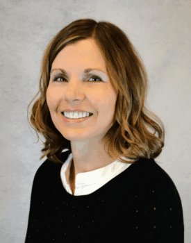 Carlen Keaster, a woman with shoulder-length light brown hair, wearing a black sweater and white collared shirt, smiles in front of a plain light gray background.