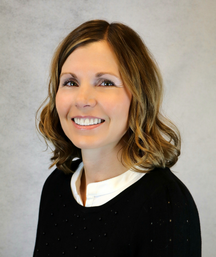 Carlen Keaster, a woman with shoulder-length light brown hair, wearing a black sweater and white collared shirt, smiles in front of a plain light gray background.