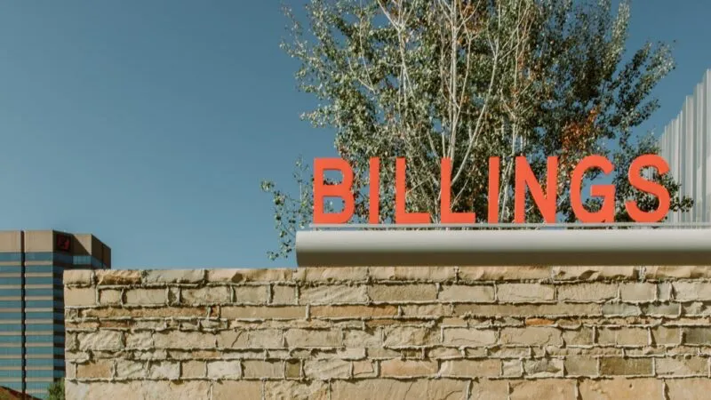 A sign with red capital letters spelling "BILLINGS" sits atop a stone wall. Trees are visible behind the wall, and a tall building is in the background against a clear blue sky.