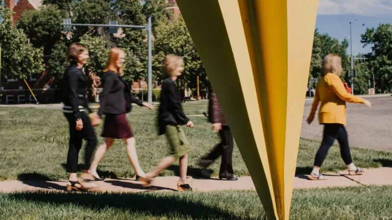 Four people walk on a sidewalk near a large yellow sculpture in a park-like area with trees and a road in the background.
