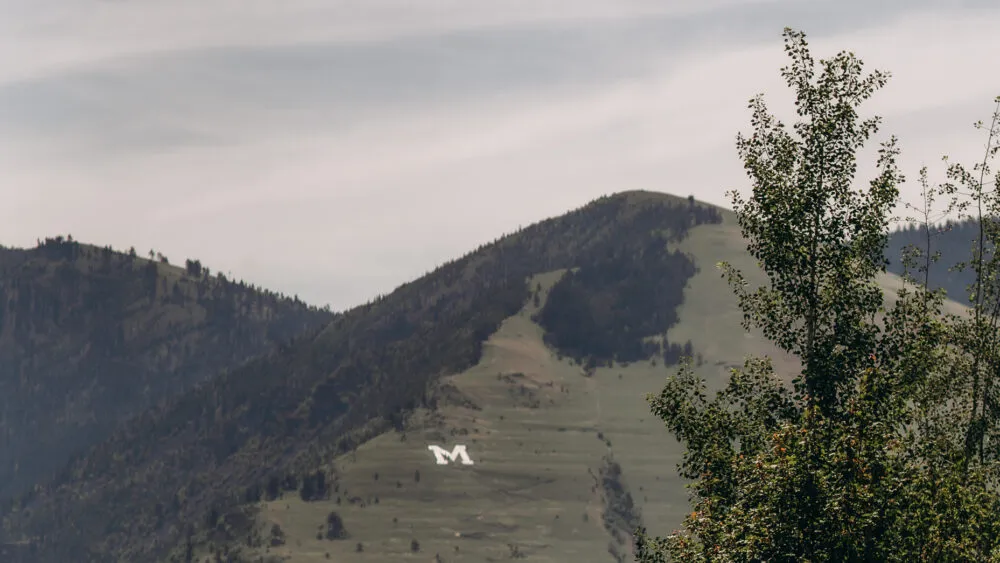 Mountain with a large white "M" on its slope, surrounded by trees and a cloudy sky.
