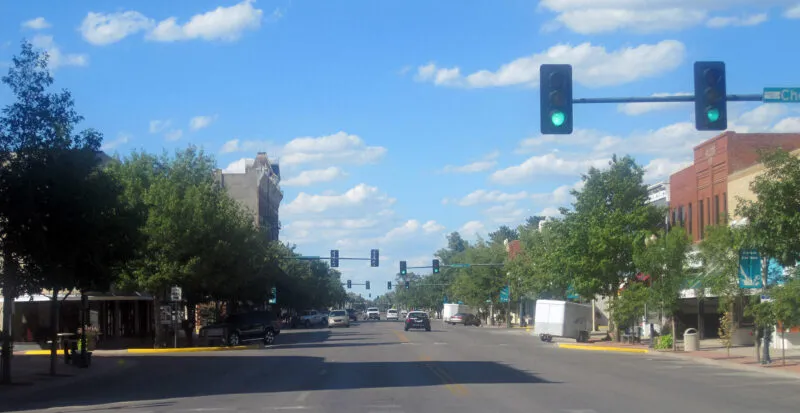 A street view of a city intersection with traffic lights, buildings, and parked cars on either side. The sky is blue with scattered clouds.