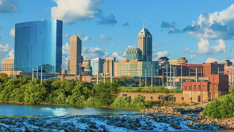 Skyline of Indianapolis with modern skyscrapers, a mix of older buildings, and a river in the foreground under a partly cloudy sky.