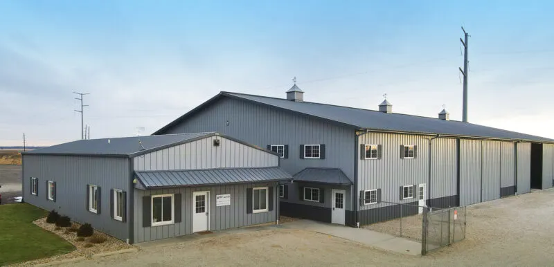 A large, gray metal warehouse with multiple garage doors and a small attached office area, surrounded by a gravel lot under a clear sky.
