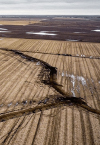 Aerial view of a landscape reveals fields with large cracks snaking through the parched soil, hinting at the need for land advisory. The terrain appears dry, with distant patches of water under an overcast sky.