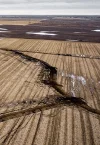 Aerial view of a landscape reveals fields with large cracks snaking through the parched soil, hinting at the need for land advisory. The terrain appears dry, with distant patches of water under an overcast sky.