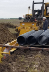 A construction worker skillfully maneuvers machinery, securing three large pipes in a muddy field. Nearby, another worker offers guidance with the support of Land Advisory expertise.