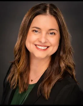 Michelle Ellis with long brown hair smiling, wearing a dark blazer and green top against a dark background, exuding confidence and professionalism, reminiscent of someone ready to excel in transactional advisory.