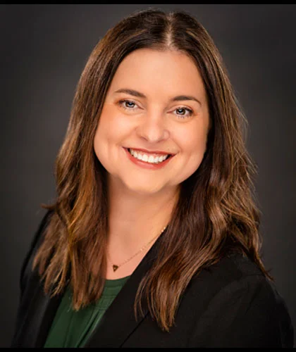 Michelle Ellis with long brown hair smiling, wearing a dark blazer and green top against a dark background, exuding confidence and professionalism, reminiscent of someone ready to excel in transactional advisory.