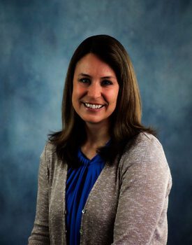 A woman with long brown hair, wearing a blue blouse and beige cardigan, poses against a blue-gray background, exuding confidence as if ready for her next transactional advisory meeting.