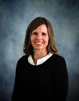 A smiling person with brown hair, clad in a black top and white collar, exudes confidence against a blue-gray background, embodying the essence of transactional advisory expertise.