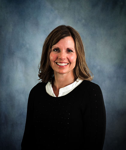 A smiling person with brown hair, clad in a black top and white collar, exudes confidence against a blue-gray background, embodying the essence of transactional advisory expertise.