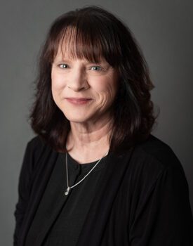 A woman with shoulder-length dark hair, donning a black sweater and necklace, smiles confidently against a gray background, embodying the essence of transactional advisory excellence.