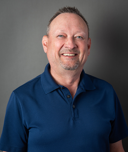 A smiling man with short hair and a goatee, embodying professional confidence, stands against a plain gray background in a blue polo shirt. His demeanor suggests expertise in transactional advisory, ready to offer insightful guidance.