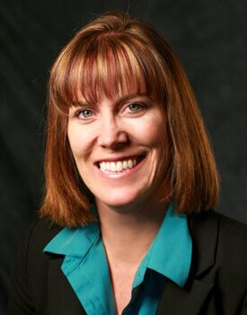 A person with shoulder-length brown hair and bangs, wearing a teal shirt and black blazer, exudes confidence while smiling against a dark background, embodying the professionalism of transactional advisory.