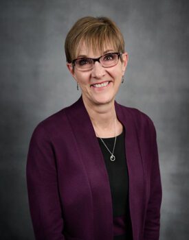 A person with short hair and glasses is smiling, exuding confidence in a dark purple cardigan over a black top. Their poised demeanor hints at their expertise in transactional advisory, set against a subtle gray background.