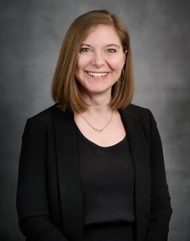 A person with shoulder-length brown hair, wearing a black blazer and top, smiles confidently against a gray background, exuding the poised professionalism often associated with transactional advisory.