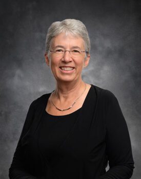 A smiling person with short gray hair and glasses, donning a black top, stands confidently against a dark gray background, embodying the essence of transactional advisory expertise.