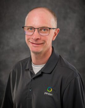 A man with glasses and a grey polo shirt featuring the Pinion logo stands confidently against a plain dark background, offering a smile that speaks to his expertise in transactional advisory.
