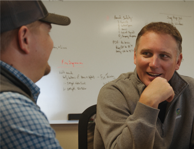 Two men in casual clothing are discussing transactional advisory strategies in an office. A whiteboard with various notes is visible in the background, hinting at their collaborative planning process.