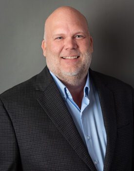 A bald man with a gray beard, exuding confidence in a dark plaid suit jacket and light blue shirt, smiles against a gray backdrop, embodying the professionalism expected in Transactional Advisory.