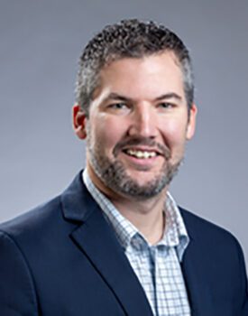 A man with short gray hair and a beard is wearing a dark suit jacket over a checkered shirt, embodying the confident charm of a transactional advisory expert. He smiles warmly at the camera against a plain gray background.