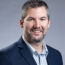 A man with short gray hair and a beard is wearing a dark suit jacket over a checkered shirt, embodying the confident charm of a transactional advisory expert. He smiles warmly at the camera against a plain gray background.